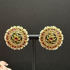 May include: A pair of gold-toned stud earrings with a circular design. Each earring features a central cluster of green and red stones surrounded by smaller stones and a border of white beads. The earrings are displayed on a black stand.
