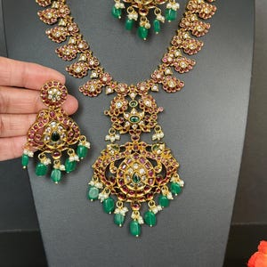 May include: A gold-toned necklace and earring set with intricate detailing. The jewelry features red and green gemstones, white pearls, and a floral design. The necklace and earrings are displayed on a black mannequin.