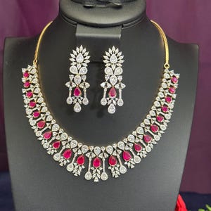 May include: A gold-toned necklace and earring set. The necklace features a series of red teardrop-shaped stones interspersed with clear, round-cut stones. The matching earrings have a similar design. The jewelry is displayed on a black mannequin.
