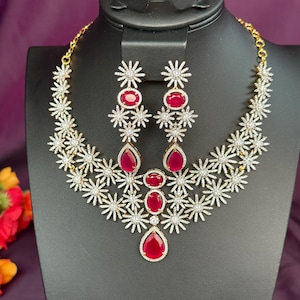 May include: A dazzling jewelry set featuring a necklace and matching earrings. The necklace and earrings are adorned with sparkling clear crystals and vibrant red gemstones. The design includes a floral pattern, creating an elegant and eye-catching accessory.