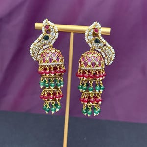May include: Gold-toned chandelier earrings with a peacock-inspired design. The earrings feature a red, green, and white beaded cascade, with a dome-shaped top adorned with red stones. The earrings are displayed on a gold stand against a purple backdrop.