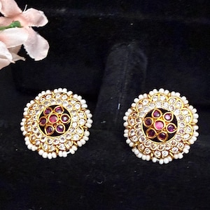 May include: A pair of gold-toned stud earrings. Each earring features a central cluster of small, round, red gemstones surrounded by clear, sparkling stones and a border of tiny white beads. The earrings are set against a dark background.