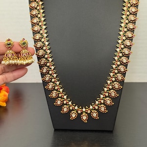 May include: A gold-toned necklace and matching earrings set. The necklace features a repeating pattern of red, green, and white gemstones. The earrings have a similar design with small white pearls. The jewelry is displayed on a black mannequin.