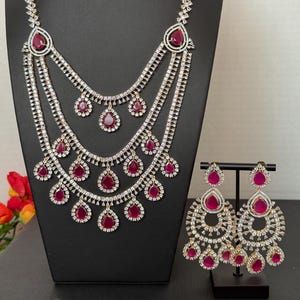 May include: A dazzling jewelry set featuring a multi-layered necklace and matching earrings. The necklace and earrings showcase a combination of clear, sparkling stones and vibrant red teardrop-shaped gems. The set is displayed on a black stand.