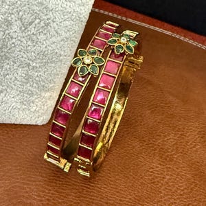 May include: A gold-toned hinged bangle bracelet featuring a row of square-cut magenta stones. The bracelet is accented with a floral motif composed of marquise-cut green stones surrounding a central clear stone, creating a vibrant contrast.