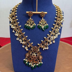 May include: A gold-toned necklace and earring set displayed on a blue bust. The necklace and earrings feature red and green gemstones, white pearls, and intricate detailing. The jewelry is designed with a floral motif.