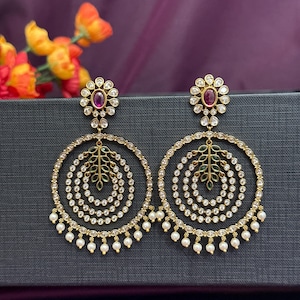 May include: A pair of gold-toned earrings with a floral design at the top and a large circular hoop. The hoops are adorned with small, clear stones and a leaf-like design. Small white pearls dangle from the bottom of the hoops. A deep purple stone is set in the center of the floral design.