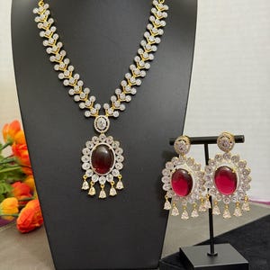 May include: A gold-toned necklace and earring set. The necklace features a central oval pendant with a deep red stone and dangling accents. The earrings mirror the pendant design. The jewelry is displayed on a black stand.