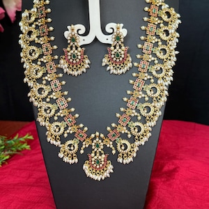 May include: A gold-toned necklace and earring set with intricate detailing. The necklace features a series of connected floral and geometric shapes, adorned with red, green, and white accents. The matching earrings echo the necklace's design, creating a cohesive and elegant jewelry set.