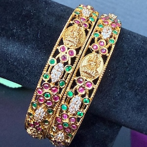 May include: Two gold bangles featuring intricate filigree metalwork, embedded with small round red and green stones, clear white crystals, and a central circular medallion depicting a deity figure seated on a lotus flower.
