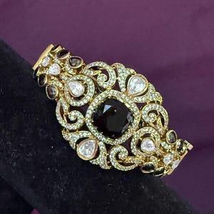 May include: Gold-toned bracelet with an ornate design. The bracelet features a large, dark gemstone at the center, surrounded by smaller clear and black gemstones. The intricate filigree work is embellished with numerous small, clear crystals.