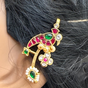 May include: A gold-toned earring with a bird-like design. The earring features red, green, and white gemstones, along with small pearls. The design includes a star, flower, and teardrop shape. The earring is worn on a person's ear.