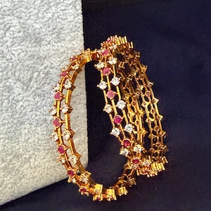 May include: Gold-toned bangles with alternating red and white gemstones. The bangles are stacked, showcasing their intricate design and sparkling details. The jewelry is displayed against a dark blue background.