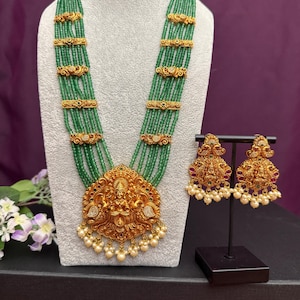May include: A gold-toned necklace and earring set. The necklace features multiple strands of green beads, gold accents, and a large pendant with a deity figure and pearl drops. The earrings are ornate, with red accents and pearl drops.
