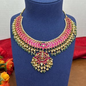 May include: A vibrant, ornate necklace with a pink and gold design. The necklace features a curved, pink upper section with a repeating pattern, accented with small pearls and green beads. A matching pendant hangs below, also in pink and gold.