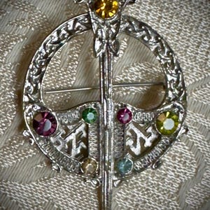 May include: Silver-toned brooch with a circular design and a central sword. The brooch features colorful gemstones in shades of yellow, purple, green, and blue. The intricate Celtic knotwork adds detail to this vintage jewelry piece.
