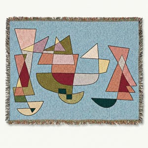 May include: A woven throw blanket with a light blue background and an abstract geometric design. The design features shapes in shades of pink, red, green, and yellow. The blanket has a fringed edge.