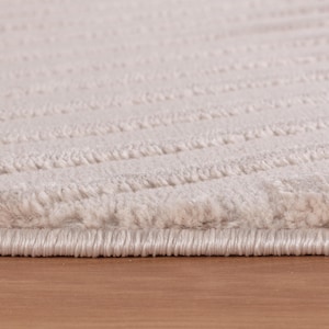 May include: Close-up of a beige rug with a textured, wavy pattern. The rug has a soft, plush appearance and is made of a thick material. The edges are neatly finished, and the rug is placed on a wooden surface.