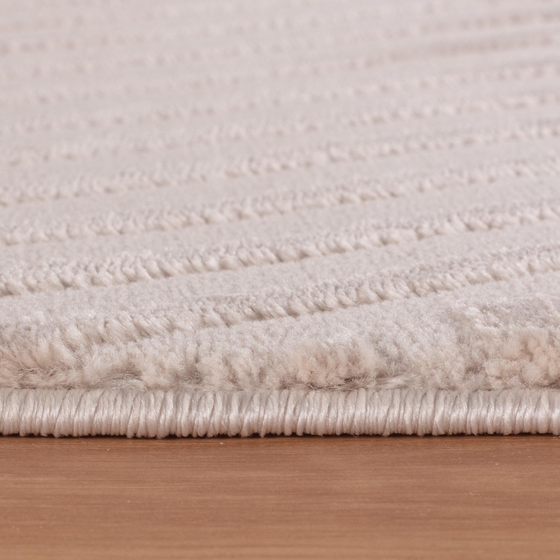 May include: Close-up of a beige rug with a textured, wavy pattern. The rug has a soft, plush appearance and is made of a thick material. The edges are neatly finished, and the rug is placed on a wooden surface.