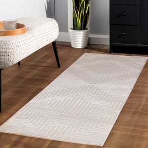 May include: A rectangular, light beige rug with a geometric zigzag pattern. The rug is placed on a hardwood floor. The rug's design features alternating light and darker beige lines, creating a textured appearance. The rug is a long, narrow runner.