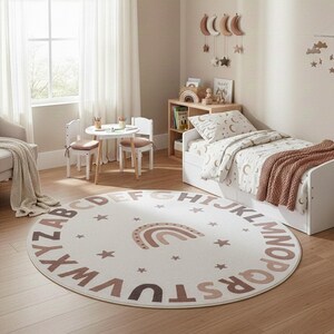 May include: A round, off-white rug with a brown alphabet border and a rainbow graphic. The rug is in a child's bedroom, alongside a bed, small table, and chairs. The room has a neutral color palette.