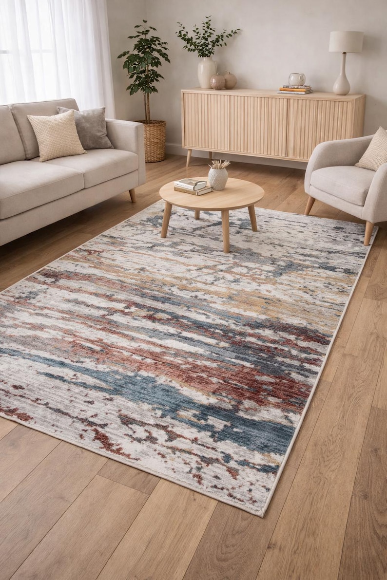 May include: A rectangular area rug with a distressed, abstract design. The rug features a blend of colors, including white, blue, red, and tan, creating a textured, painterly effect. It is placed on a hardwood floor in a living room setting.