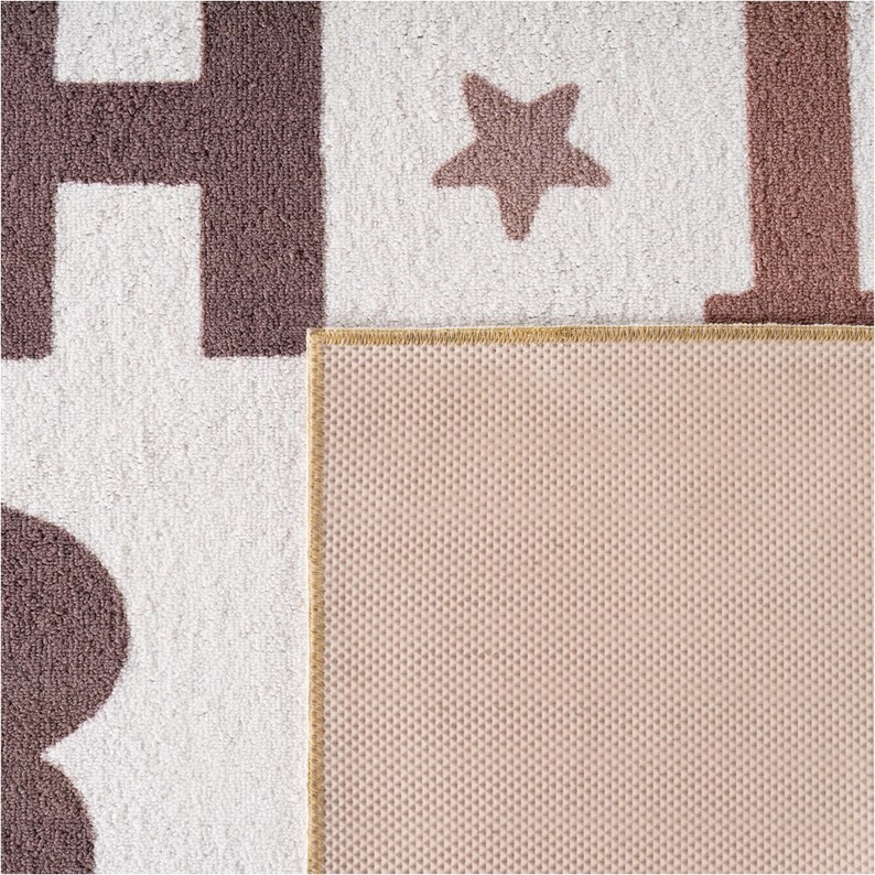 May include: A close-up of a white and brown rug with large letters and a star design. The rug has a textured surface and a tan border. The back of the rug is a light pink color.