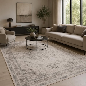 Könnte beinhalten: Ein rechteckiger Teppich mit einem abgenutzten, vintage-inspirierten Muster in Creme-, Beige- und Anthrazittönen. Das Design umfasst ein zentrales Medaillon-Motiv, das von einer komplizierten floralen Bordüre umgeben ist und den Boden bedeckt.