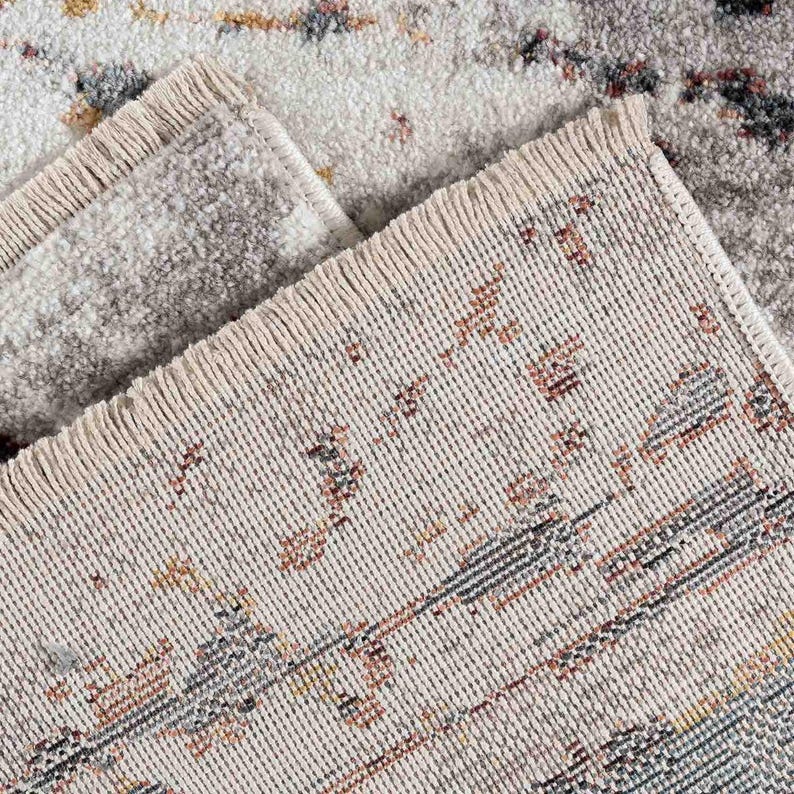 May include: Close-up of a beige area rug with a distressed pattern. The rug features a mix of colors, including gray, brown, and yellow, creating a vintage look. The edges have a fringed detail.