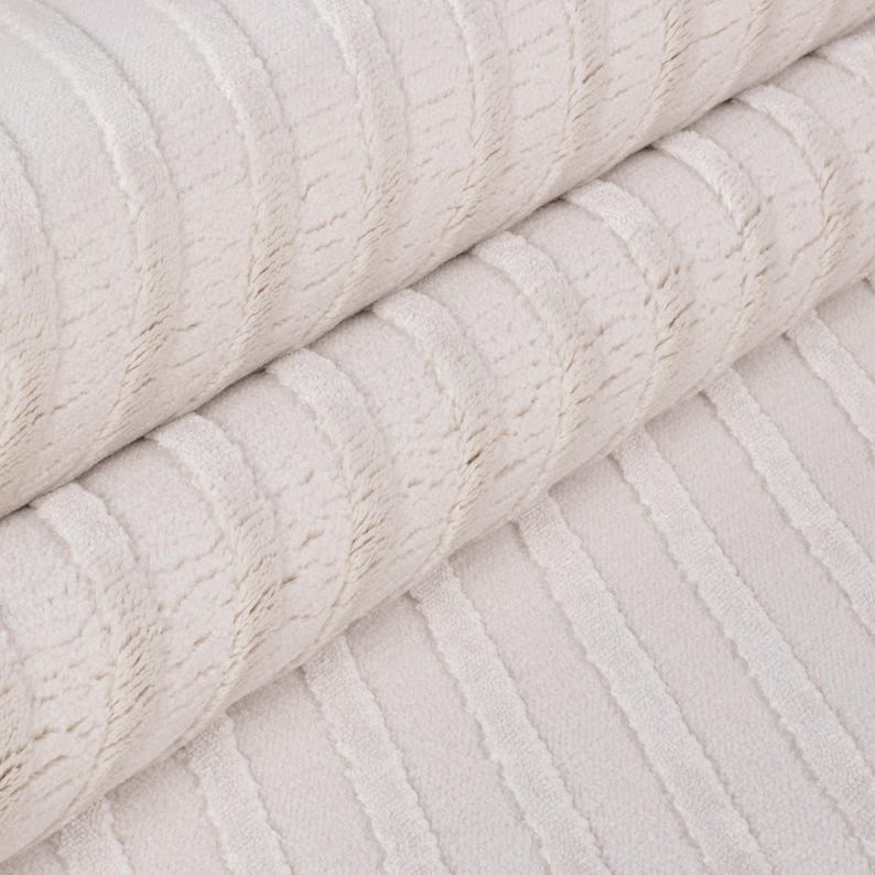 May include: Close-up of a rolled-up, off-white fabric with a textured, ribbed pattern. The fabric appears soft and plush, with vertical lines creating a subtle visual texture. The neutral color suggests a versatile textile.