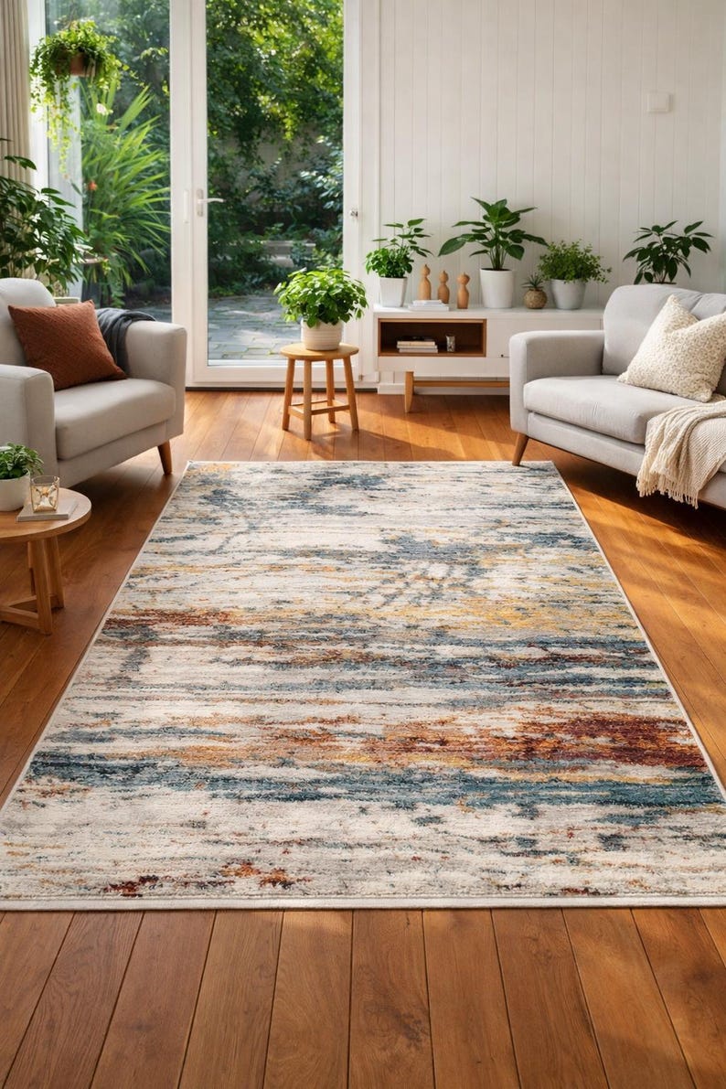 May include: A rectangular area rug with a distressed, abstract design. The rug features a blend of colors including cream, blue, gold, and rust. It is placed on a hardwood floor in a living room setting, with neutral-toned furniture and plants.