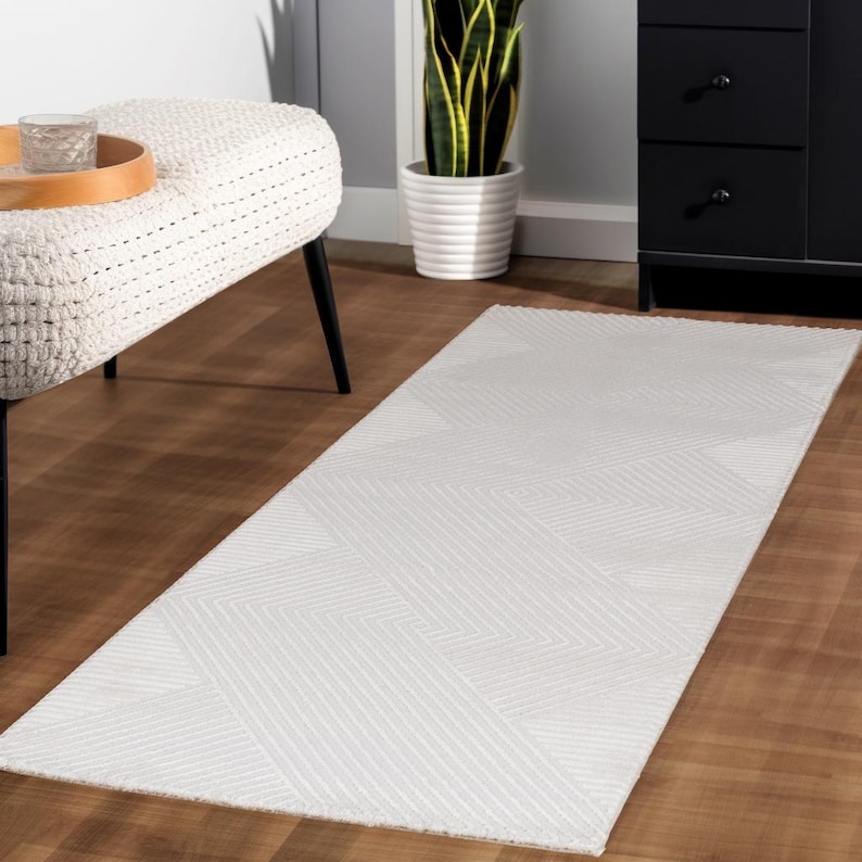 May include: A rectangular white rug with a geometric pattern of angled lines. The rug is placed on a wooden floor, and the design creates a textured appearance. The rug is a neutral color and could be used in a variety of interior design settings.