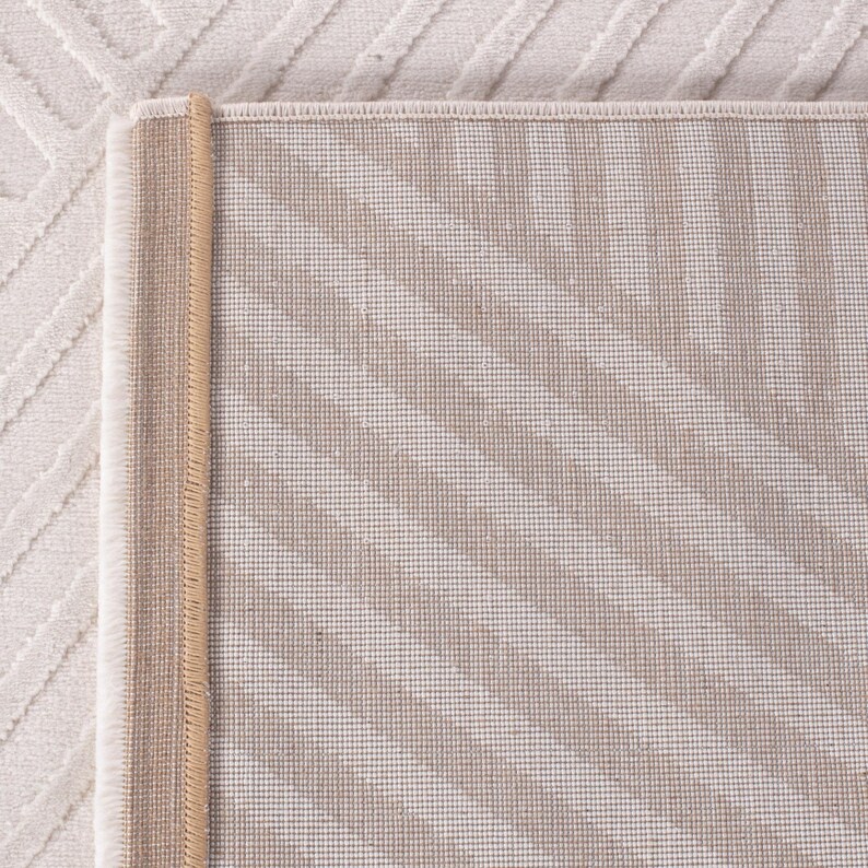 May include: Close-up of a beige and white rug with a geometric pattern. The rug has a textured surface with a diagonal striped design in beige and white. The edge is finished with a tan border.