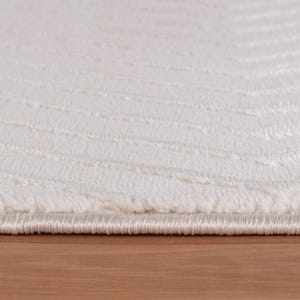 May include: Close-up of a white rug with a textured, striped pattern. The rug has a soft, plush appearance and a subtle sheen. The edge of the rug is visible, showing a finished border. The rug is placed on a wooden surface.