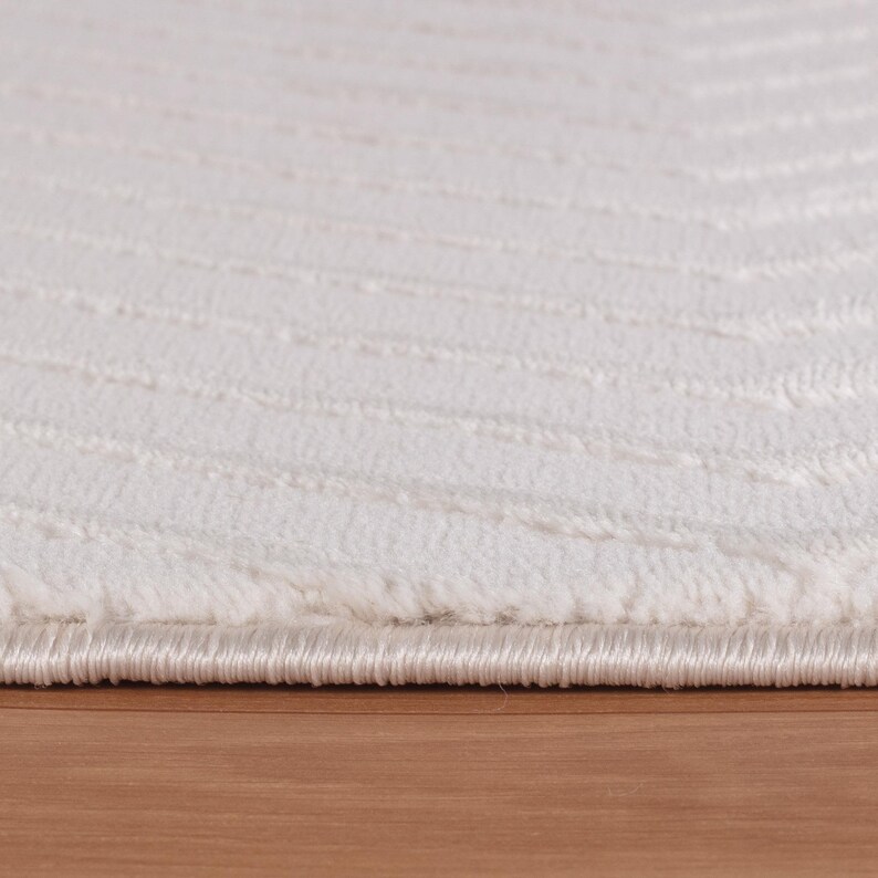 May include: Close-up of a white rug with a textured, striped pattern. The rug has a soft, plush appearance and a subtle sheen. The edge of the rug is visible, showing a finished border. The rug is placed on a wooden surface.