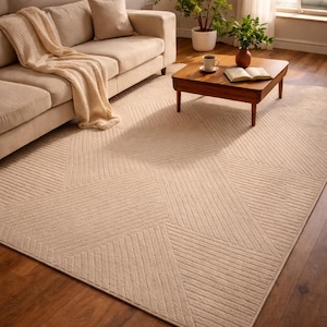May include: Beige geometric patterned area rug with a textured surface. The rug features a repeating design of angled lines, creating a modern aesthetic. The rug is placed on a hardwood floor, with a sofa and coffee table in the background.