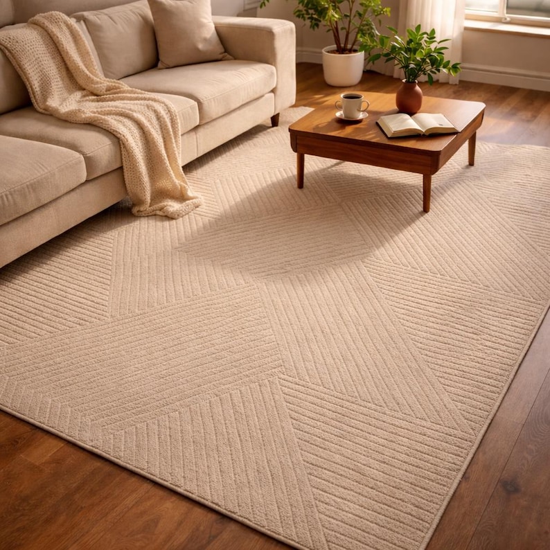 May include: Beige geometric patterned area rug with a textured surface. The rug features a repeating design of angled lines, creating a modern aesthetic. The rug is placed on a hardwood floor, with a sofa and coffee table in the background.
