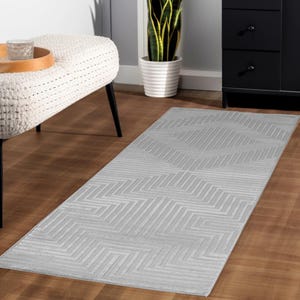 May include: A rectangular, light gray rug with a geometric pattern of angled lines. The rug is placed on a hardwood floor, and the design creates a textured appearance. The rug is a decorative element for home decor.