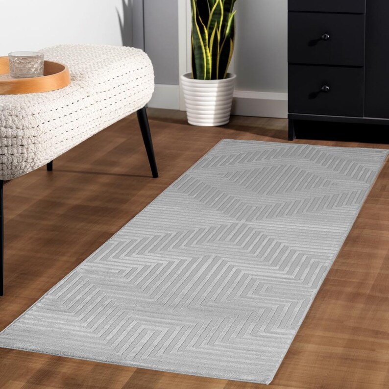 May include: A rectangular, light gray rug with a geometric pattern of angled lines. The rug is placed on a hardwood floor, and the design creates a textured appearance. The rug is a decorative element for home decor.