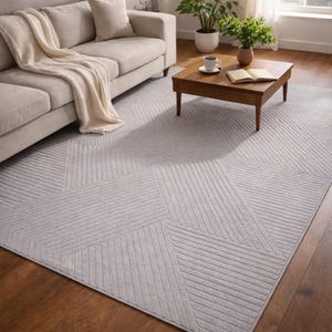 May include: A light gray geometric patterned area rug with a textured surface. The rug features a design of angled lines and shapes. A beige sofa with a cream-colored throw blanket is in the background.