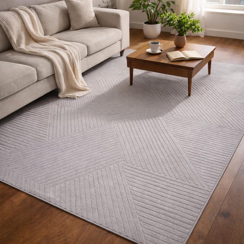 May include: A light gray geometric patterned area rug with a textured surface. The rug features a design of angled lines and shapes. A beige sofa with a cream-colored throw blanket is in the background.