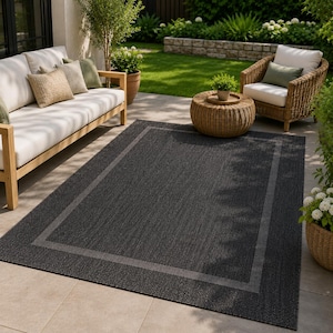 Peut inclure: Un tapis d'extérieur rectangulaire présentant un centre gris anthracite foncé avec une bordure gris plus clair. Le tapis possède une texture tissée et est placé sur le sol d'une terrasse devant un canapé clair et un fauteuil en osier.