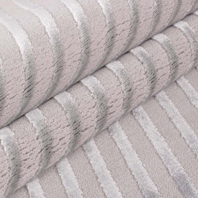 May include: Close-up of a rolled-up, light gray fabric with vertical, textured stripes in a lighter shade of gray. The fabric appears soft and plush, with a subtle sheen. The stripes create a sense of depth and dimension.