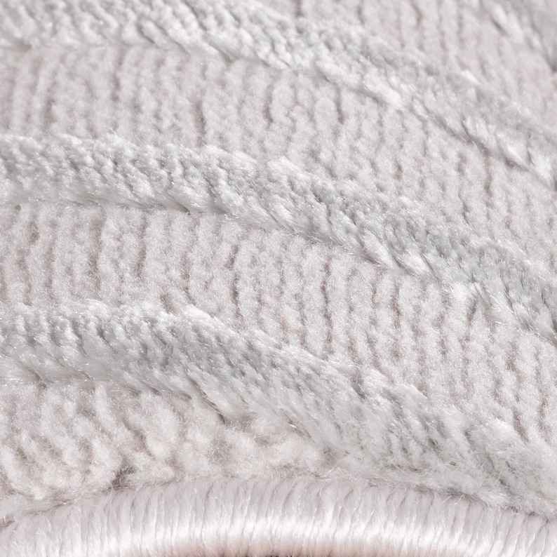 May include: Close-up of a soft, textured rug in shades of white and light gray. The rug features a pattern of diagonal, raised lines, creating a plush and inviting appearance. The fibers appear dense and luxurious, suggesting comfort and warmth.