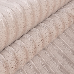 May include: Close-up of a soft, beige fabric with a ribbed texture. The fabric is folded over, revealing the detailed pattern of vertical ridges. The material appears plush and inviting, suitable for textiles.