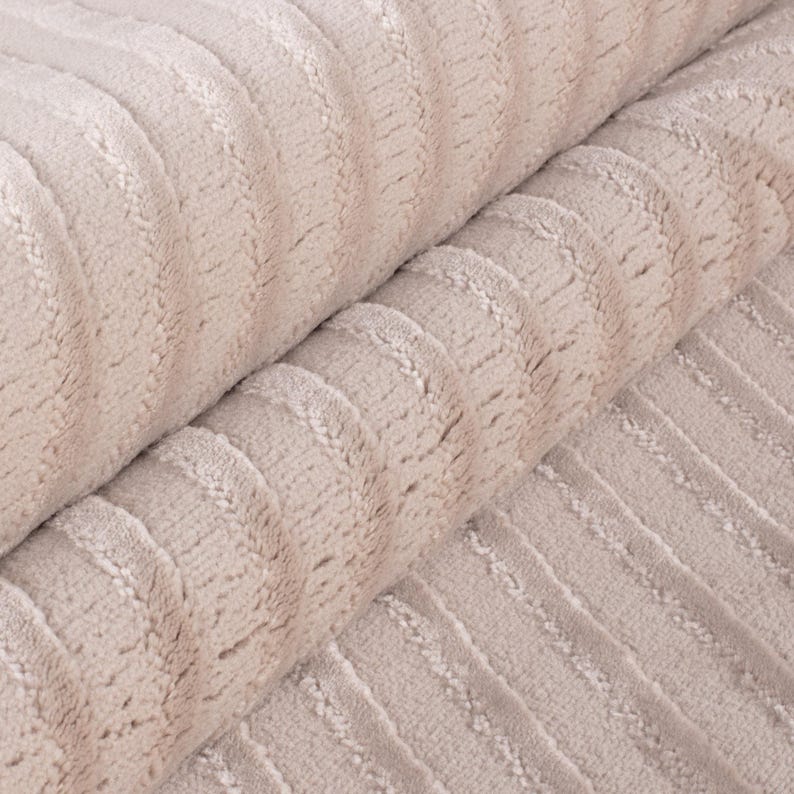 May include: Close-up of a soft, beige fabric with a ribbed texture. The fabric is folded over, revealing the detailed pattern of vertical ridges. The material appears plush and inviting, suitable for textiles.