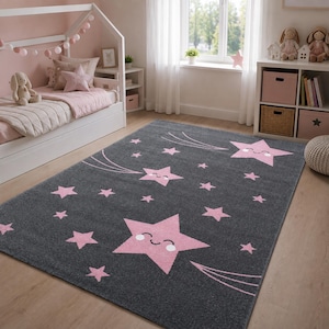 Peut inclure: Un tapis rectangulaire au fond gris foncé orné d'un motif d'étoiles rose clair. Plusieurs grandes étoiles présentent un visage souriant avec des yeux fermés et une bouche courbée, accompagnées de lignes suggérant un mouvement.