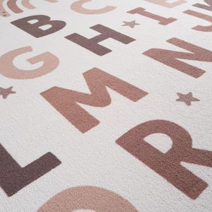 May include: A soft, off-white rug featuring a repeating alphabet design in shades of brown and tan. The letters are large and stylized, with a textured appearance. Small star shapes are scattered throughout the design. The rug has a neutral color palette.