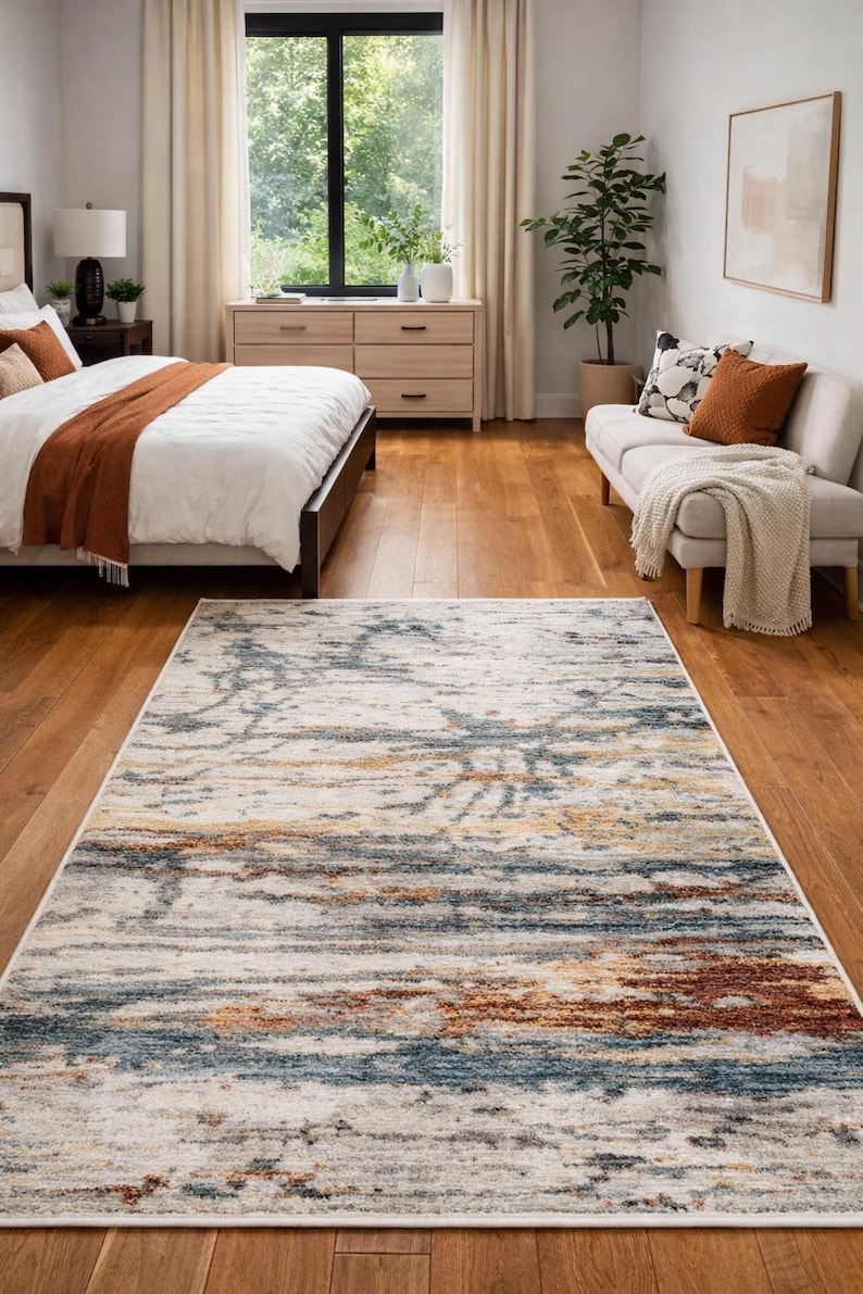 May include: A rectangular area rug with a distressed design. The rug features a mix of colors, including blue, brown, and cream, creating a textured, abstract pattern. It is placed on a hardwood floor in a bedroom setting.