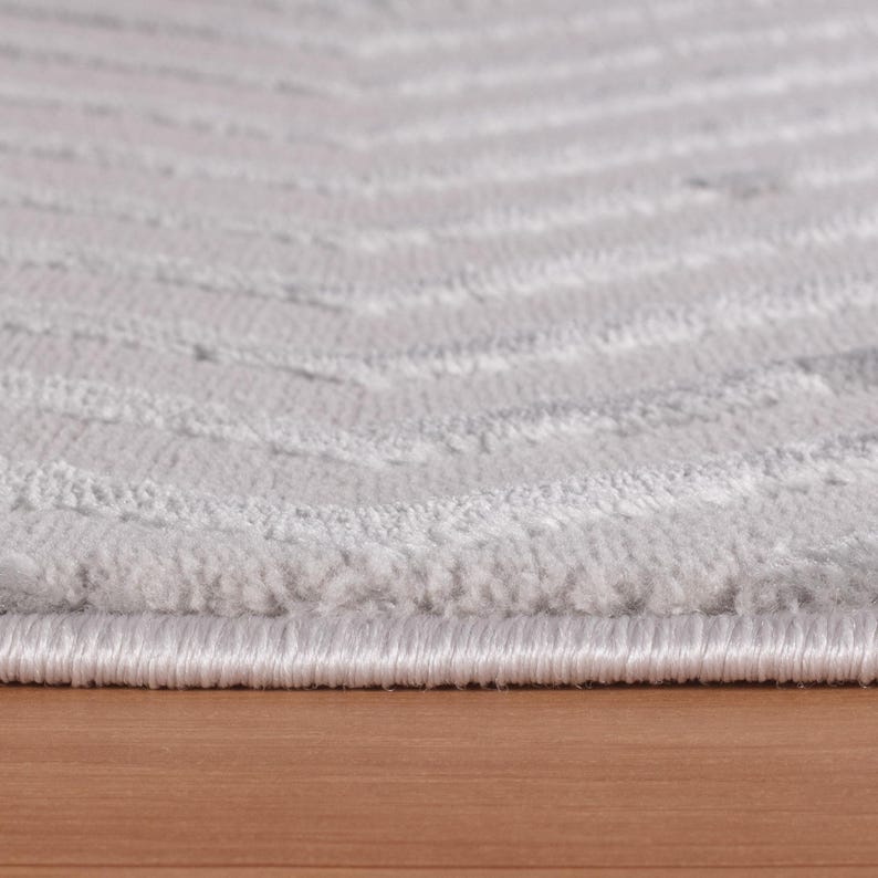 May include: Close-up of a light gray rug with a chevron pattern. The rug has a soft, textured surface with horizontal lines. The edges are finished with a white border. The rug is on a wooden surface.
